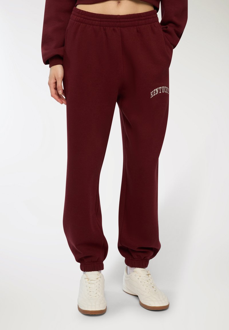 Burgundowe spodnie dresowe z elastycznym pasem i ściągaczami, z haftem "KENTUCKY" w kolorze białym. Połączone z białymi sneakersami.