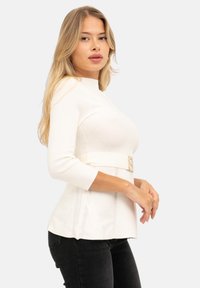 Top bianco aderente con scollatura alta, maniche a tre quarti e cintura in vita con fibbia rettangolare; abbinato a pantaloni neri.
