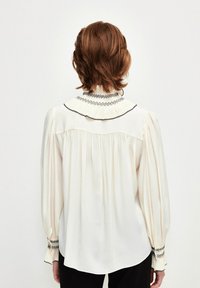 Cremefarbene Bluse mit langen Puffärmeln, einem plissierten Kragen und schwarzem Zickzackstich am Ausschnitt und an den Bündchen. Glatter, leichter Stoff.