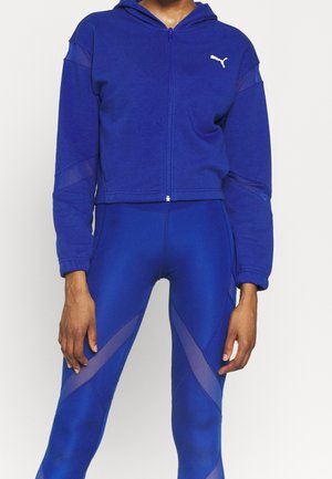 Kobieta ubrana w niebieską bluzę Puma zapinaną na zamek oraz dopasowane niebieskie legginsy z siateczkowymi panelami na rękawach i nogawkach.