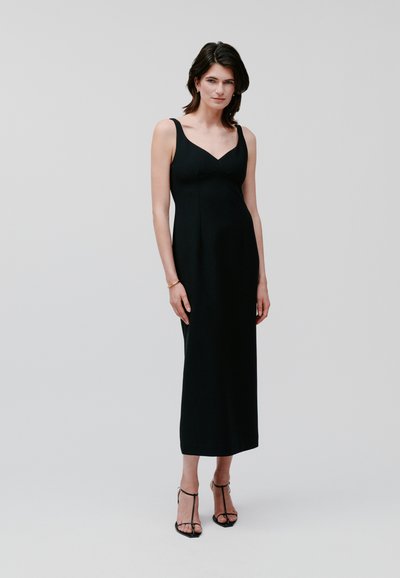 Robe maxi noire sans manches avec un corsage ajusté et un décolleté en V. Tissu doux avec une texture lisse, associée à des sandales à talons noires à lanières.