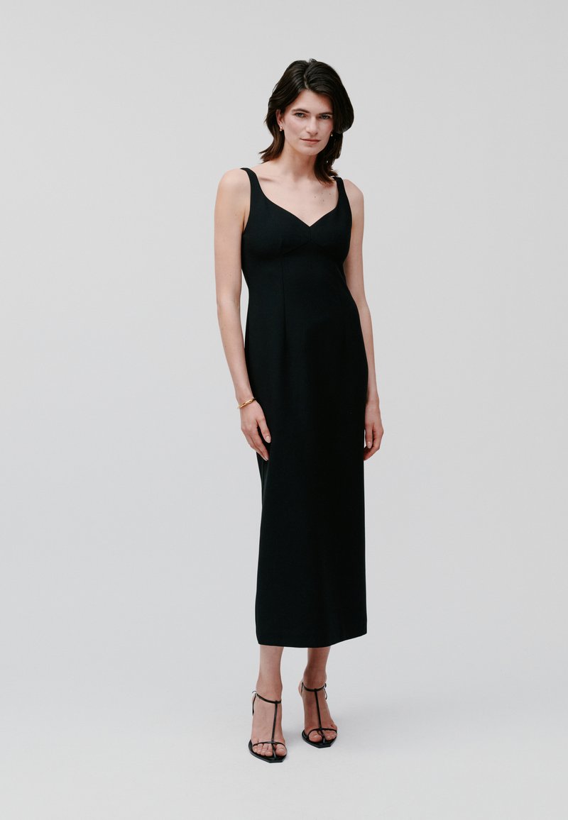 Black sleeveless maxi dress with a fitted bodice and v-neckline. Soft fabric with a smooth texture, paired with black strappy heeled sandals.