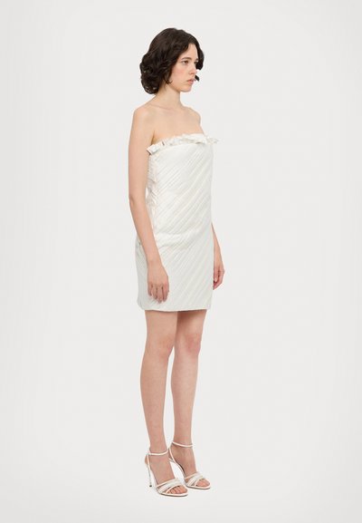 Vestido mini sin tirantes en crema pálido, con plisados verticales, un borde superior con volantes y una silueta ajustada. Combinado con tacones blancos con tiras.