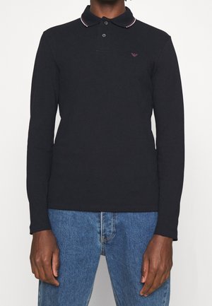 Man wearing a black long-sleeve polo shirt with a small logo on the left chest paired with blue jeans against a white background.