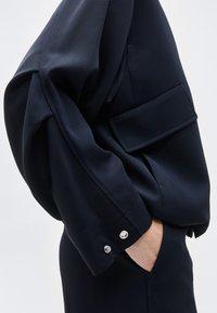 Σκούρο ναυτικό μπλε σακάκι με πλαϊνή τσέπη με καπάκι και κουμπιά στις μανσέτες, φορεμένο πάνω από ταιριαστό παντελόνι, χέρι βαλμένο στην τσέπη, σε απλό ανοιχτό φόντο.