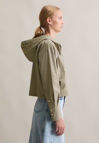 Gekürztes Kapuzenshirt im grün-beigen Karomuster, mit knöpfen in den Bündchen und lockerer Passform, kombiniert mit einem hellblauen Jeansrock.