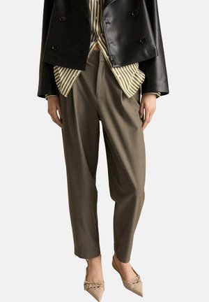 Femme portant un pantalon fuselé olive, des ballerines beiges pointues cloutées, une chemise rayée et une veste courte en cuir noir sur un fond uni.