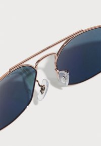 Ray-Ban'i prillid roosa kulla metallraamiga, siniste toonitud läätsede ja selgete nina padjakesega. Nurka üleva disain koos peenikeste kõrvarõngastega.