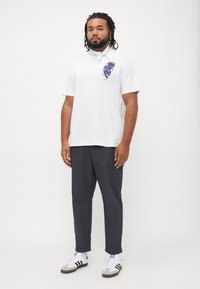 Hvid bomulds polo skjorte med et farverigt grafisk design på brystet, parret med mørkegrå bukser og hvide sneakers med sorte detaljer.