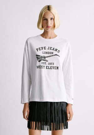 Mujer rubia que lleva una sudadera blanca de Pepe Jeans London con un estampado negro de guitarra y una falda negra con flecos, mirando hacia adelante sobre un fondo blanco.