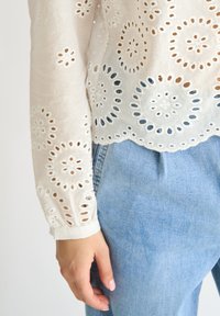 Weiße bestickte Bluse mit runden Ausschnitten und gewelltem Saum, kombiniert mit hellblauem Denim. Der Stoff ist leicht und strukturiert.
