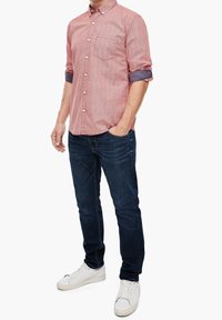 Camisa de botones con un patrón rojo y blanco, con las mangas enrolladas, combinada con unos vaqueros azul oscuro y zapatillas blancas. Diseño sencillo y casual.