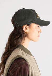 Woman in profile wearing a dark green baseball cap, gold hoop earrings, a beige quilted vest, and a dark plaid shirt against a white background.