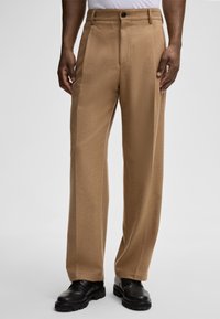 Brązowe szerokie spodnie wykonane z miękkiego materiału, z zapięciem na guzik, minimalistyczny design i gładka faktura. Noszone z czarnymi butami.