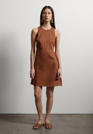 Frau mit langen dunklen Haaren, die ein ärmelloses braunes Kleid, passende braune Pantoletten und lange Quastenohrringe trägt, steht auf einem Betonboden.