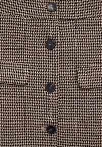 Tissu à motif pied-de-poule marron et noir avec deux poches avant et boutons noirs ; design structuré, texture lisse.