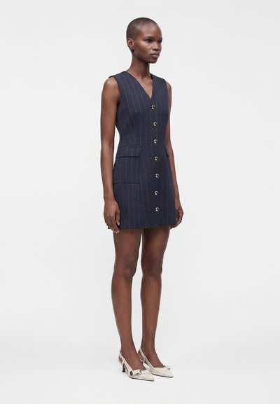 Femme portant une robe sans manches bleu marine à fines rayures, boutonnée devant, avec des poches à rabat et des talons slingback blancs à bout pointu.