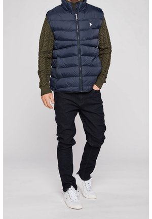 Mann trägt marineblaue gesteppte Weste über olivgrünem Zopfpullover, schwarze Hose und weiße Turnschuhe, steht mit einer Hand in der Tasche.