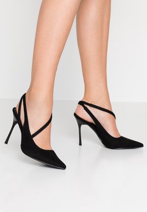 Jambes portant des chaussures à talons aiguilles noires à bout pointu avec des brides croisées fines sur une surface blanche unie.