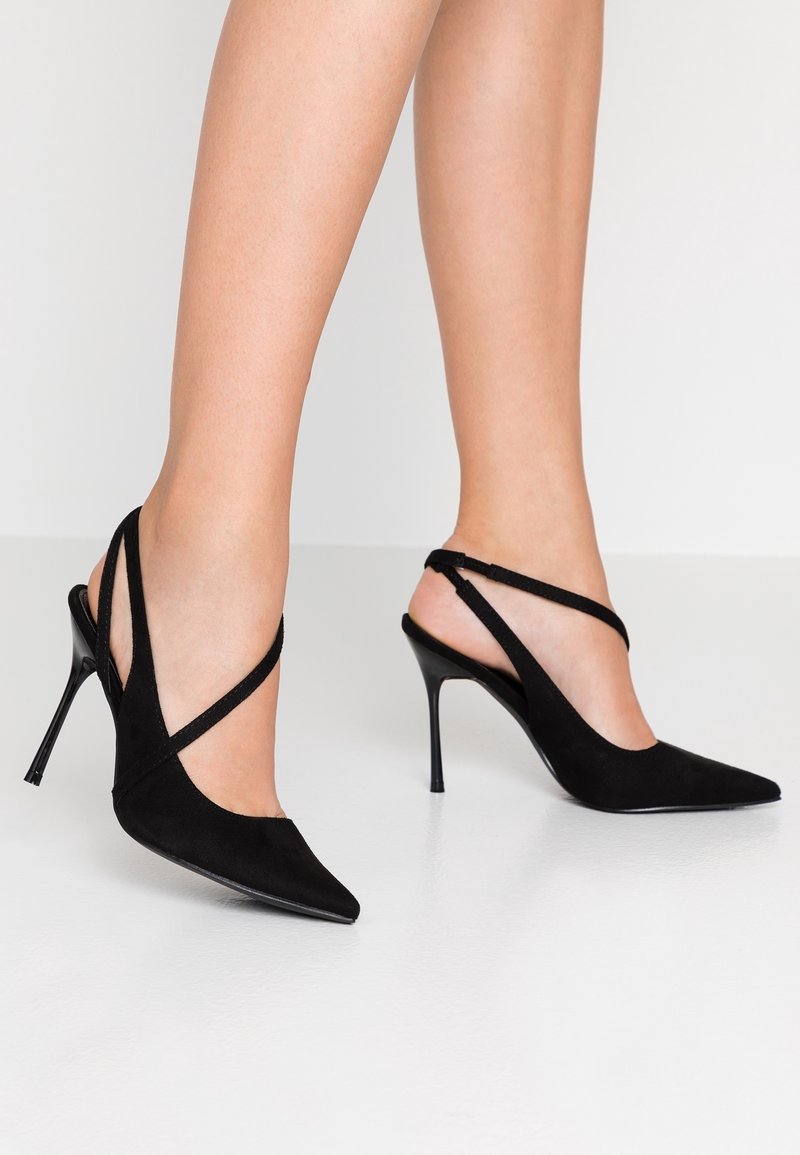 Jambes portant des chaussures à talons aiguilles noires à bout pointu avec des brides croisées fines sur une surface blanche unie.