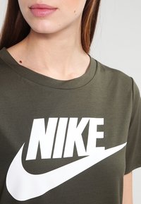Olivgrön t-shirt med rund halsringning som har en stor vit Nike-logotyp tryckt på framsidan. Slät bomullstyg.