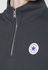 Sudadera gris de cremallera hasta el cuarto con una textura suave. Presenta un distintivo circular blanco con el logo rojo y azul de 'Converse All Star'.