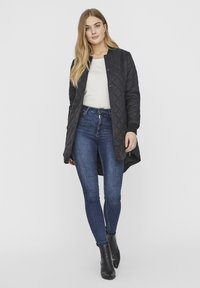 Manteau long noir matelassé avec des poignets côtelés, porté sur un haut crème et un jean foncé ajusté, associé à des bottes à chevilles noires. Fond uni.