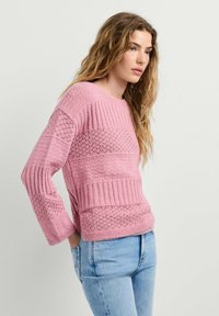 Donna con lunghi capelli ondulati che indossa un maglione rosa lavorato a maglia con trama e jeans azzurri chiari, in piedi con le mani nelle tasche posteriori, guarda avanti.