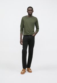 Ilgomis rankovėmis alyvuogių žalios spalvos marškiniai, juodos tiesios kojų džinsai ir smėlio spalvos sportiniai bateliai su tekstūriniais akcentais. Paprastas dizainas, be raštų ar matomų prekių ženklų.