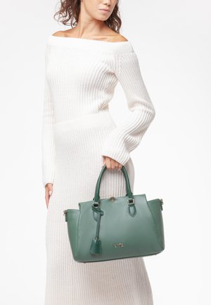 Donna con vestito bianco lavorato a maglia con spalle scoperte che tiene una grande borsa verde in pelle con doppio manico e dettagli dorati.