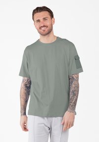 Uśmiechnięty mężczyzna z brodą i tatuażami na całych rękach nosi szaro-niebieską koszulę z krótkim rękawem i kieszeniami oraz jasne spodnie na tle białego tła.