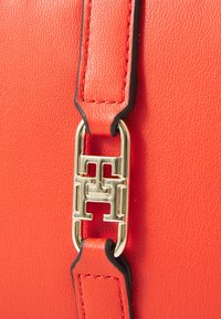 Red leather handbag detail featuring a gold-tone hardware clasp with a geometric pattern and black edge accents on the strap.