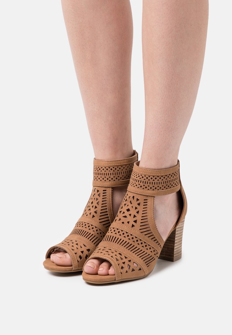 Sandalias de tacón abiertas y marrones con recortes geométricos, tira en el tobillo y un tacón de bloque texturizado. Material de piel sintética, diseño minimalista.