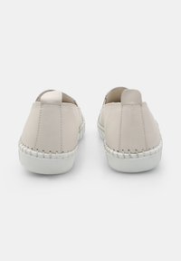 Világos bézs színű, slip-on stílusú cipők sima bőr külsőrésszel, fehér gumitalppal és díszítő varrással az oldalán. Lekerekített hátsó kialakítás.