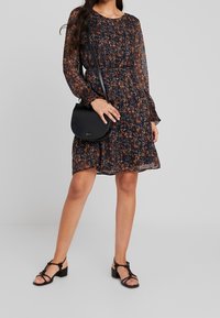 Tmavé květinové šaty s dlouhými rukávy, zdobené oranžovými a modrými vzory. V kombinaci s černou kabelkou přes rameno a černými sandály na blokovém podpatku.