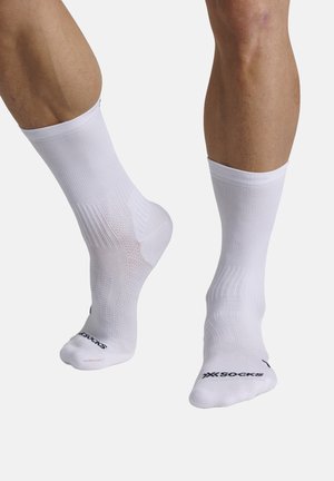 Benen met witte sportsokken tot halverwege de kuit met zwarte logotekst bij de tenen, één voet plat op de grond en de andere lichtjes op de tenen.