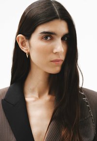 Pendientes dorados con un diseño puntiagudo, que lleva una modelo con un blazer marrón a rayas, que presenta un cuello negro en contraste.