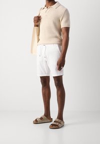 Polo shirt lavorato a maglia beige, pantaloni corti bianchi e sandali a scivolo marroni. L'outfit presenta una vestibilità ampia e include una borsa tote beige.