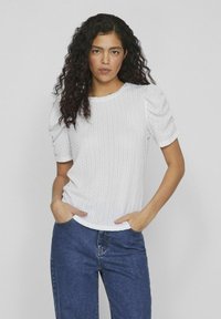 Haut blanc texturé à manches courtes avec des manches bouffantes, col rond, associé à un jean en denim bleu. Présente un motif de tissu subtil.