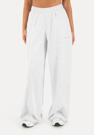 Persona que lleva pantalones deportivos sueltos de color gris claro con cintura elástica y zapatillas blancas, mostrada desde la cintura hasta los pies sobre un fondo liso.