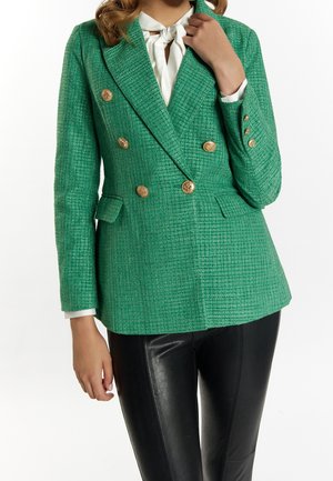Femme portant un blazer vert texturé à double boutonnage avec des boutons dorés, un chemisier blanc avec foulard et un pantalon en cuir noir.
