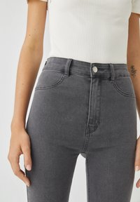 Personne portant un jean gris taille haute et un débardeur blanc c ôtelé, montrant les mains et un petit tatouage floral sur le bras gauche.