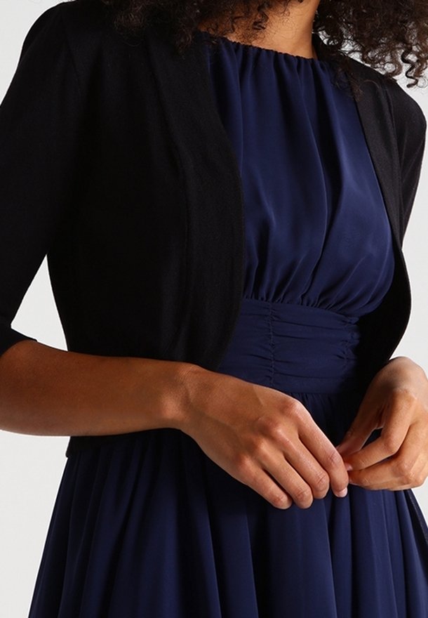 Mujer vestida con un vestido azul marino recogido, con una cinturilla fruncida y un cárdigan abierto negro de tres cuartos de manga.