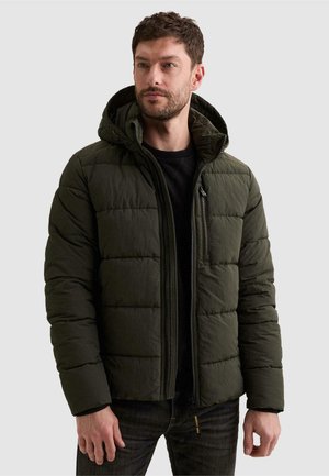 Homme portant une doudoune matelassée vert foncé avec capuche par-dessus une chemise noire, debout devant un fond clair uni.