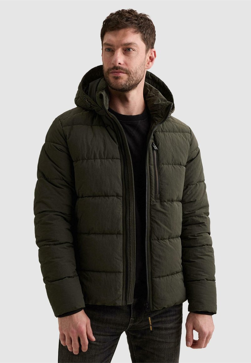 Homme portant une doudoune matelassée vert foncé avec capuche par-dessus une chemise noire, debout devant un fond clair uni.