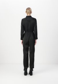 Zwart jumpsuit met lange mouwen, elastische taille en aansluitende manchetten. Opvallende details zijn een kraag en een knoopsluiting bij de pols.