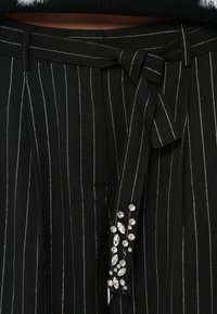 Pantalon rayé noir avec ceinture en tissu, présentant des lignes verticales argentées et des strass décoratifs sur le côté.