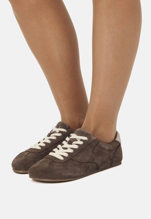 Zapatillas de ante marrón con cordones blancos, un diseño de perfil bajo y una textura suave. Presentan un acento en el talón de contraste y detalles de costura sutiles.