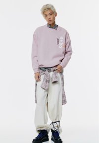 Felpa oversized rosa chiaro con logo, camicia grigia a strati, pantaloni cream larghi e stivali scuri con lacci blu. Camicia a quadri legata in vita.