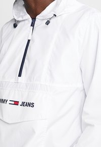 Biały polarowy kurtka z kapturem, z przednią kieszenią oraz granatowymi akcentami. Na kieszeni nadrukowane logo marki "TOMMY JEANS".